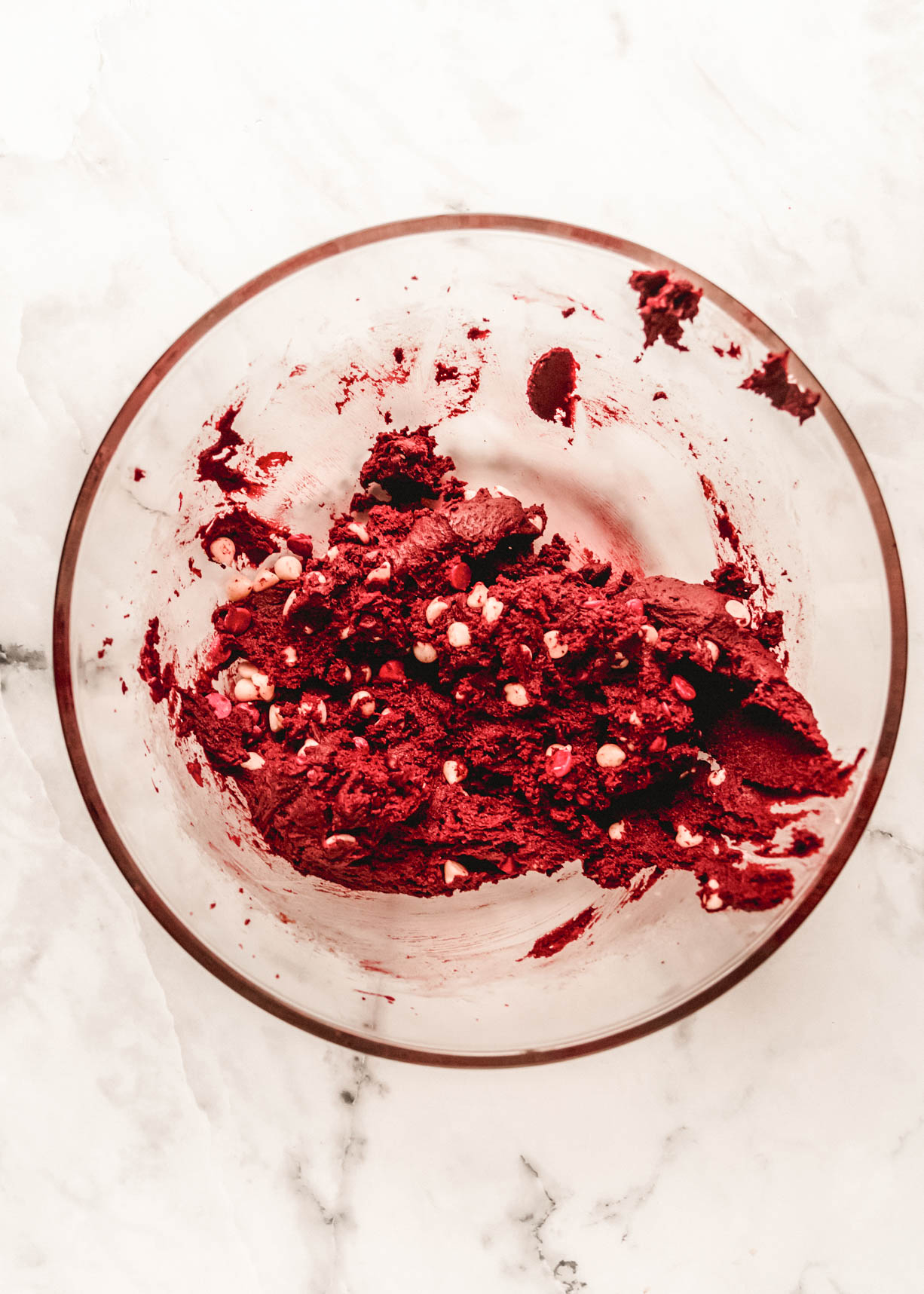 Overhead view of red velvet chocolate chip cookie dough mixed with white chocolate chips in a clear glass bowl on a light marble surface.