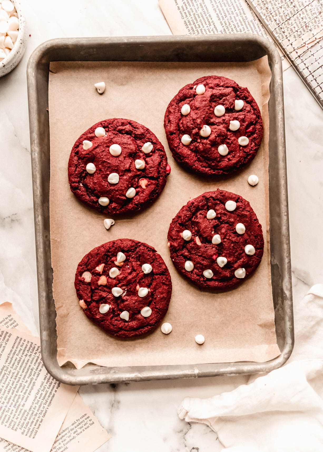 Overhead view of chewy red velvet chocolate chip cookies baked on a parchment-lined metal baking sheet, topped with white chocolate chips and styled on a light marble surface.
