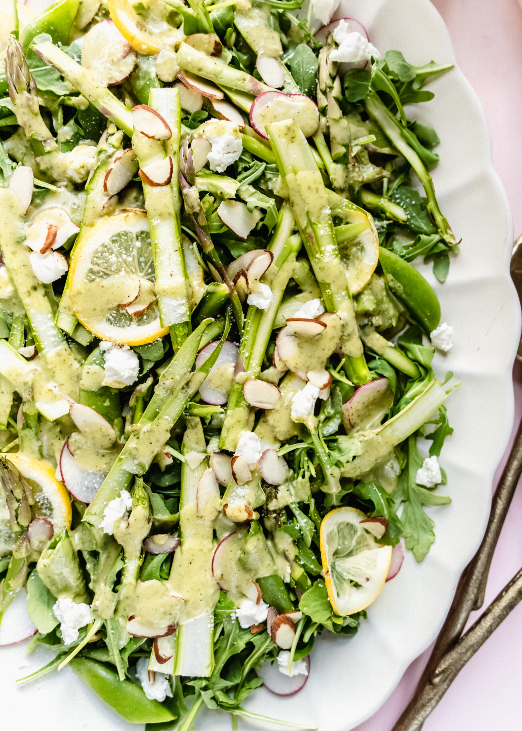 Close-up of a raw asparagus salad with shaved asparagus ribbons, arugula, sugar snap peas, thinly sliced radishes, crumbled goat cheese, and toasted almonds, drizzled with creamy lemon basil dressing and garnished with fresh lemon slices on a white platter.