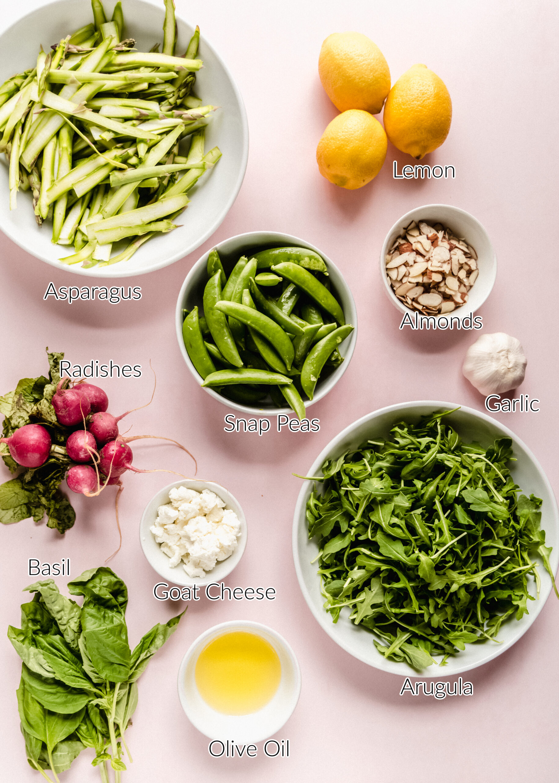 Overhead view of ingredients for a raw asparagus salad arranged on a pink background, including shaved asparagus ribbons, fresh arugula, sugar snap peas, sliced almonds, goat cheese, radishes, basil, garlic, lemons, and olive oil in small bowls.