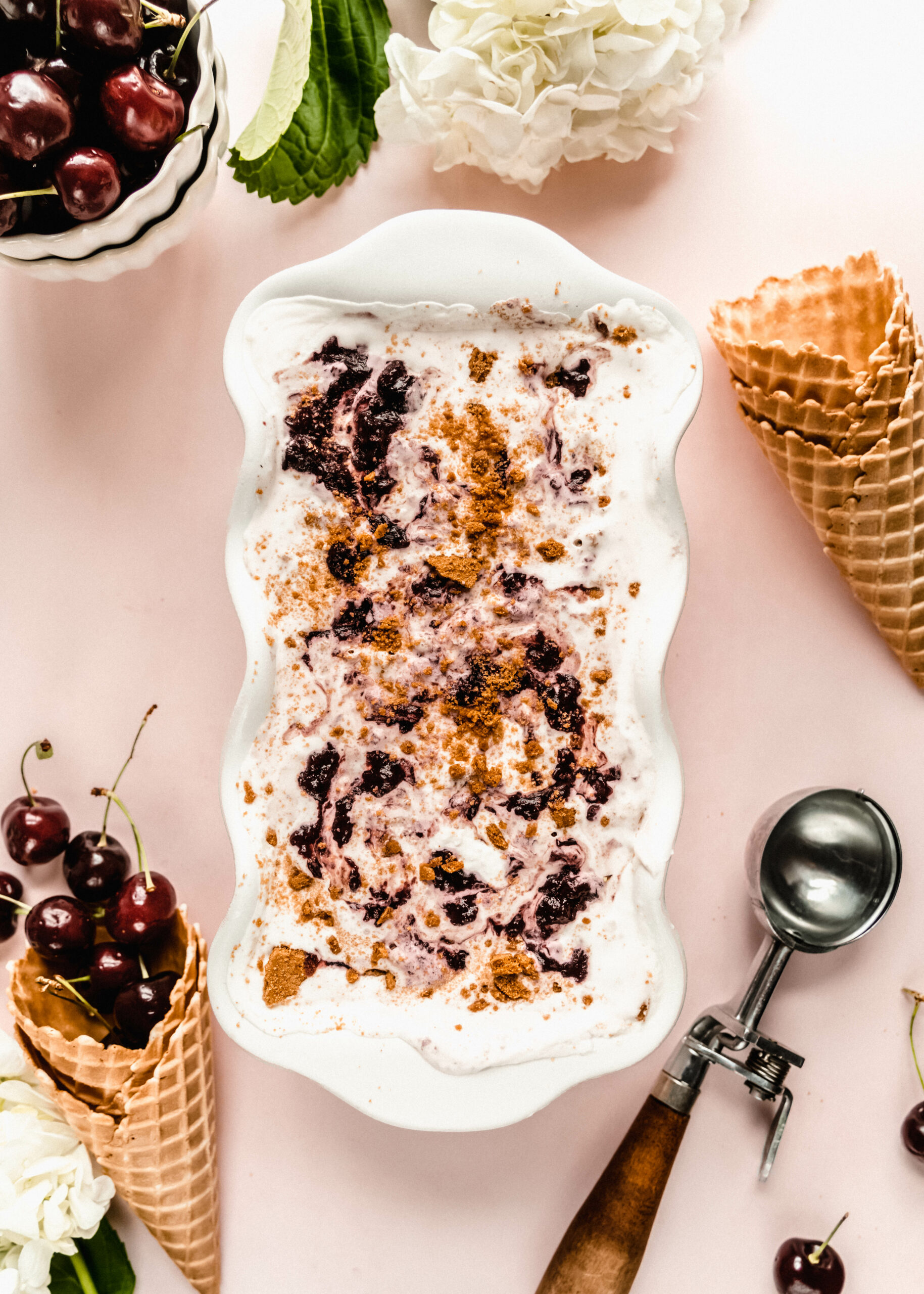 cherry cheesecake ice cream before being scooped in white loaf pan, surrounded by waffle cones and an ice cream scoop