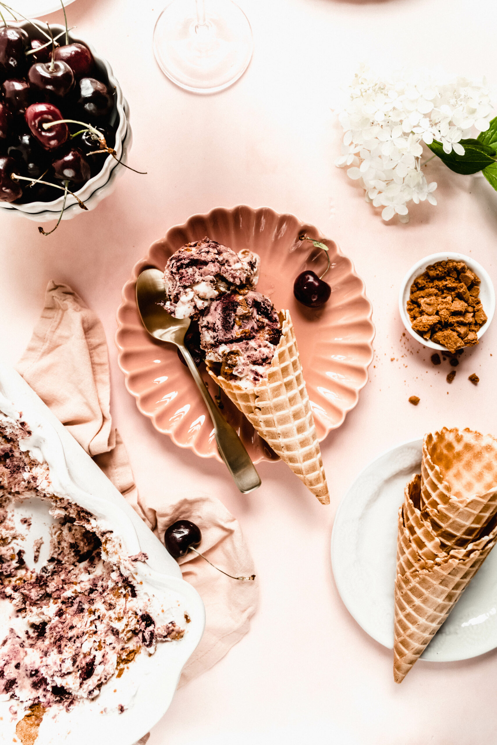 cherry cheesecake ice cream scooped in waffle cone on pink scalloped plate, surrounded by cherries in a bowl, white hydrangeas and pink napkin on a pink background.