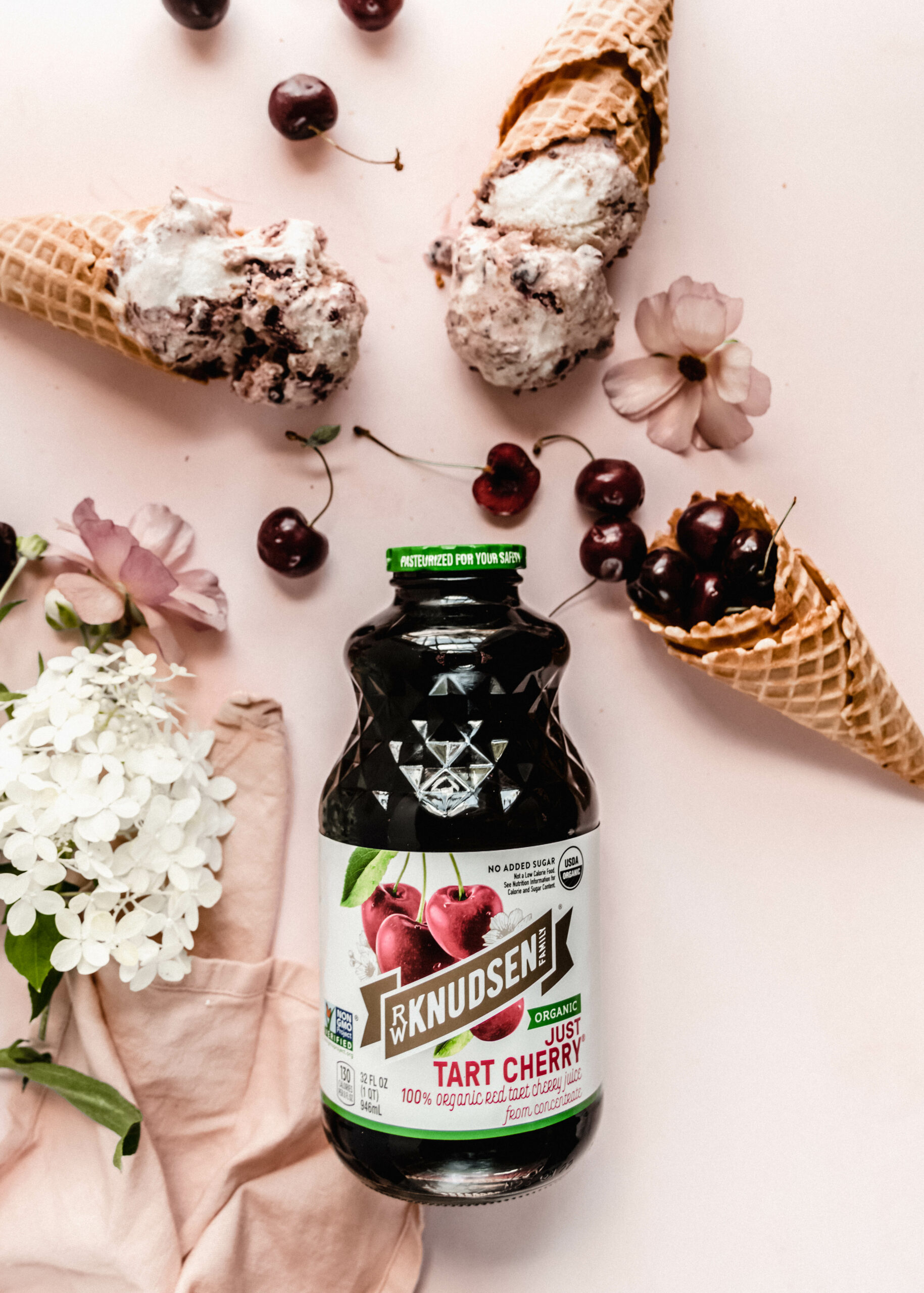 cherry juice bottle on pink background surrounded by cherry cheesecake ice cream in waffle cones