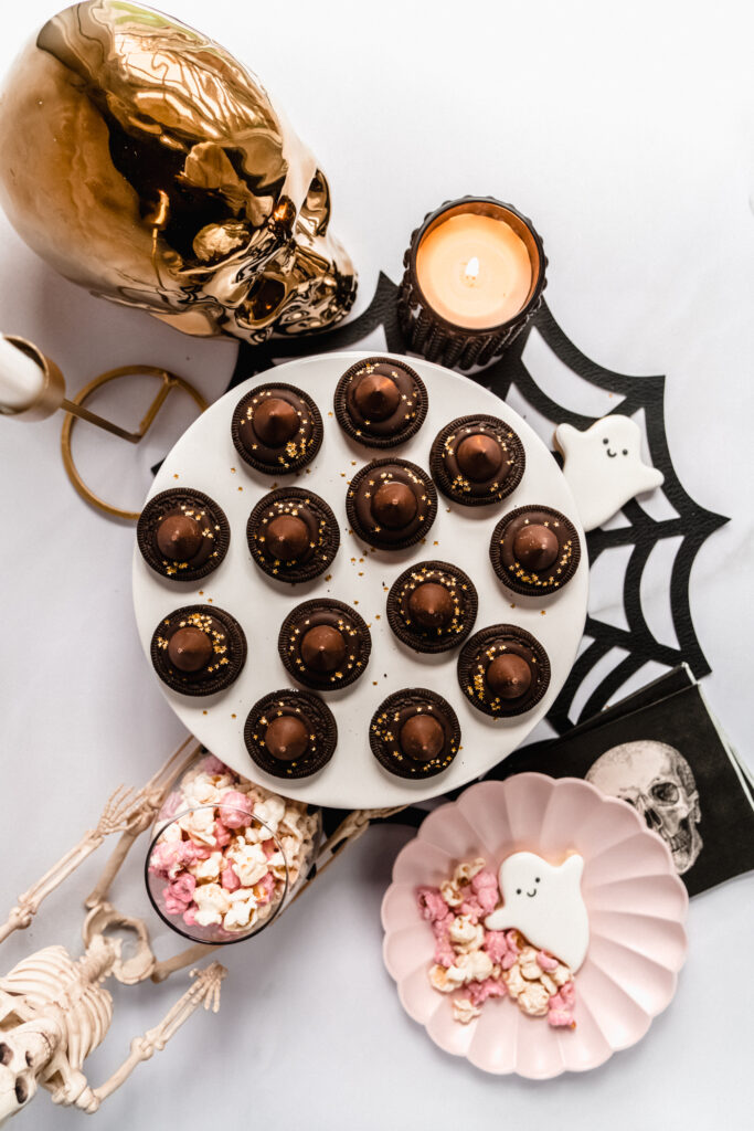 overhead shot of Halloween witch hat cookies on a white cake plate placed on a black spider web place mat, surrounded by ghost cookies, a gold skull and festive halloween popcorn.