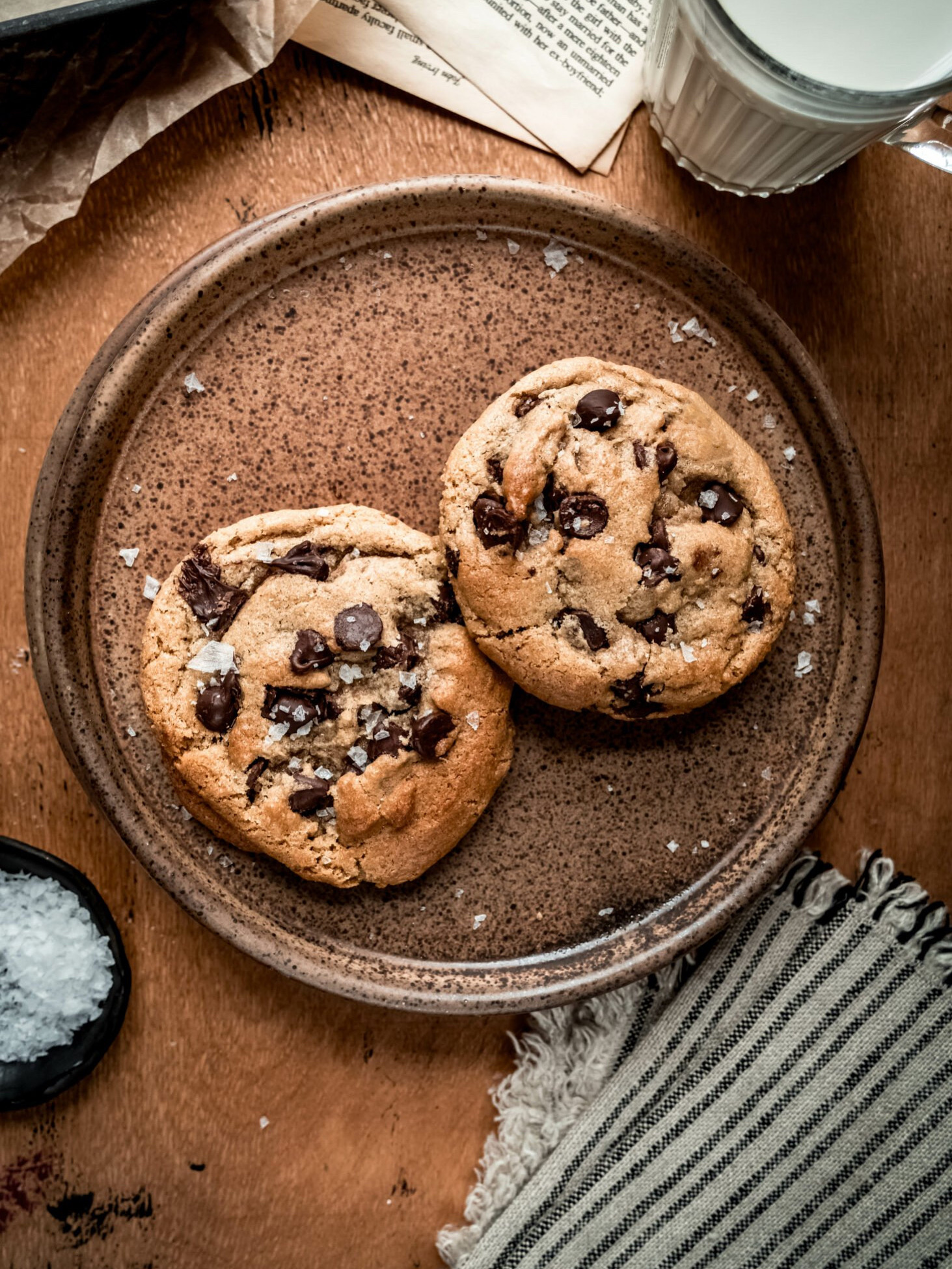 Alt text: Two of the best chocolate chip cookies topped with flaky sea salt sit on a rustic brown ceramic plate, accompanied by a glass of milk, a pinch bowl of salt, and a striped napkin on a wooden table.