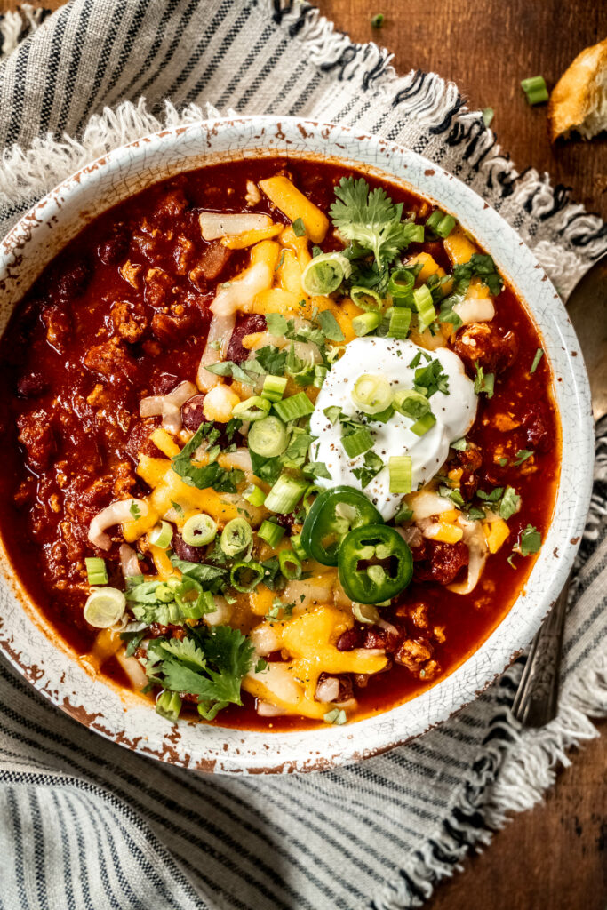 bowl of turkey chili garnished with shredded cheese, cilantro, sliced green onion and sour cream. The bowl is sitting on a striped napkin on a wood table.