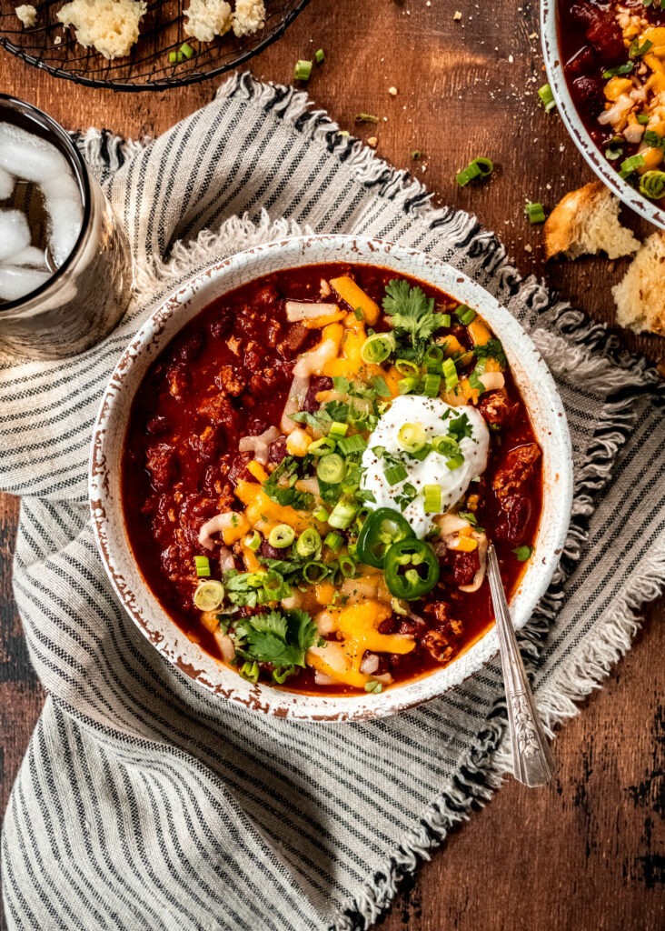 bowl of turkey chili garnished with shredded cheese, cilantro, sliced green onion and sour cream. The bowl is sitting on a striped napkin on a wood table next to a water glass.