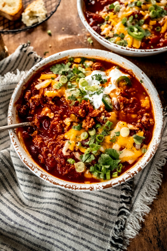 bowl of turkey chili garnished with shredded cheese, cilantro, sliced green onion and sour cream. The bowl is sitting on a striped napkin on a wood table