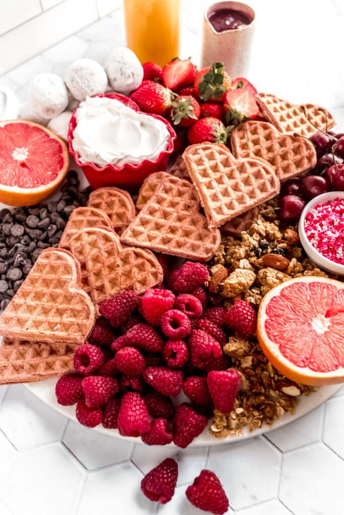 Side shot of valentines day breakfast board with heart shaped waffles, raspberries, strawberries, grapefruit, chocolate chips, granola, and shipped cream