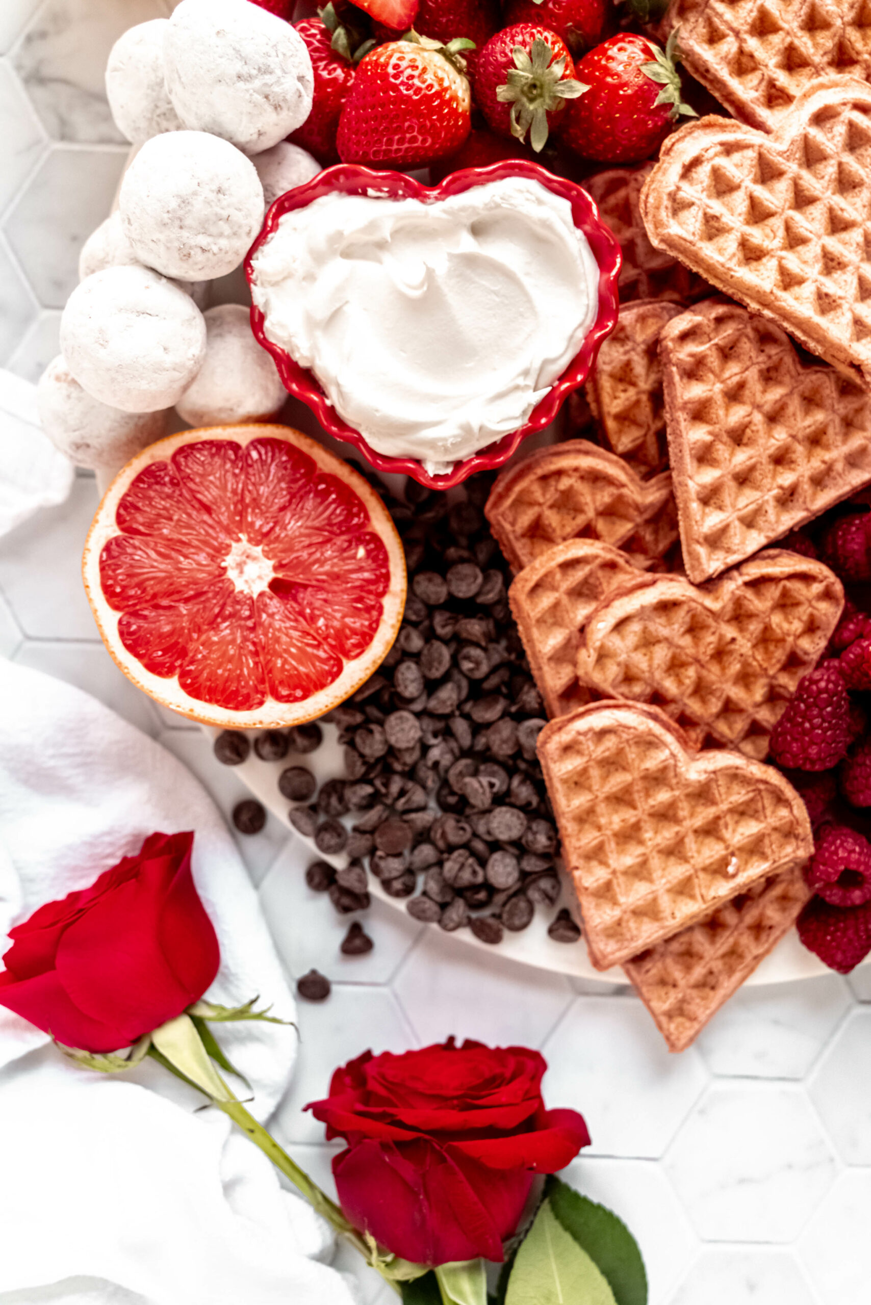 close up of whipped cream in red heart shaped ramekin on valentines day breakfast board with heart shaped waffles, raspberries, strawberries, grapefruit, chocolate chips, granola, and shipped cream