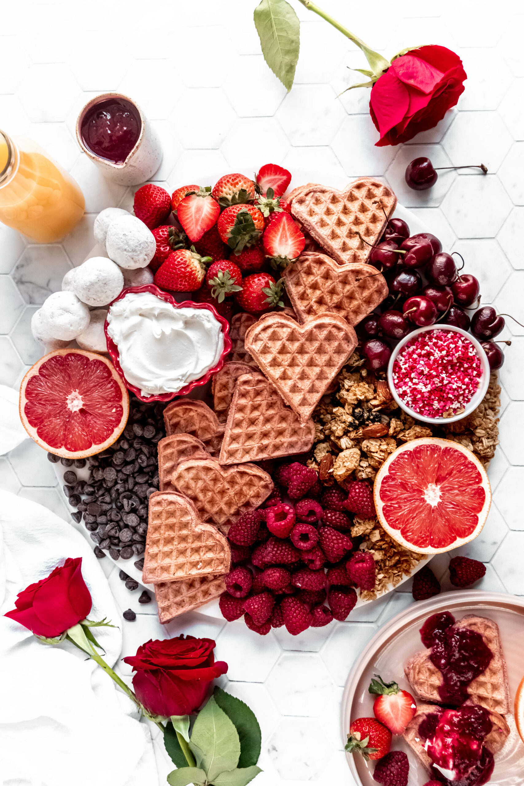 full overhead shot of valentines day breakfast board with heart shaped waffles, raspberries, strawberries, grapefruit, chocolate chips, granola, and shipped cream on hexagon marble background with red roses on the counter