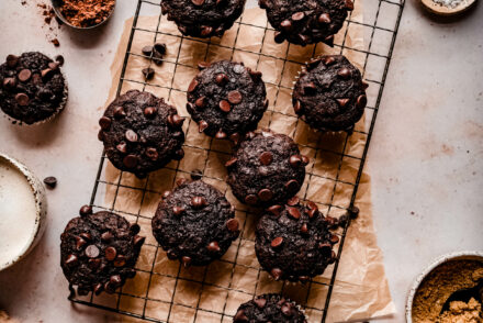 banana chocolate chip muffins on a wire cooling rack surrounded by chocolate chips, a bowl of cocoa powder and neutral linens