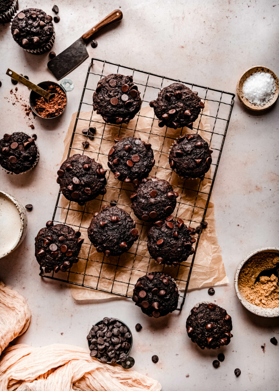 banana chocolate chip muffins on a wire cooling rack surrounded by chocolate chips, a bowl of cocoa powder and neutral linens
