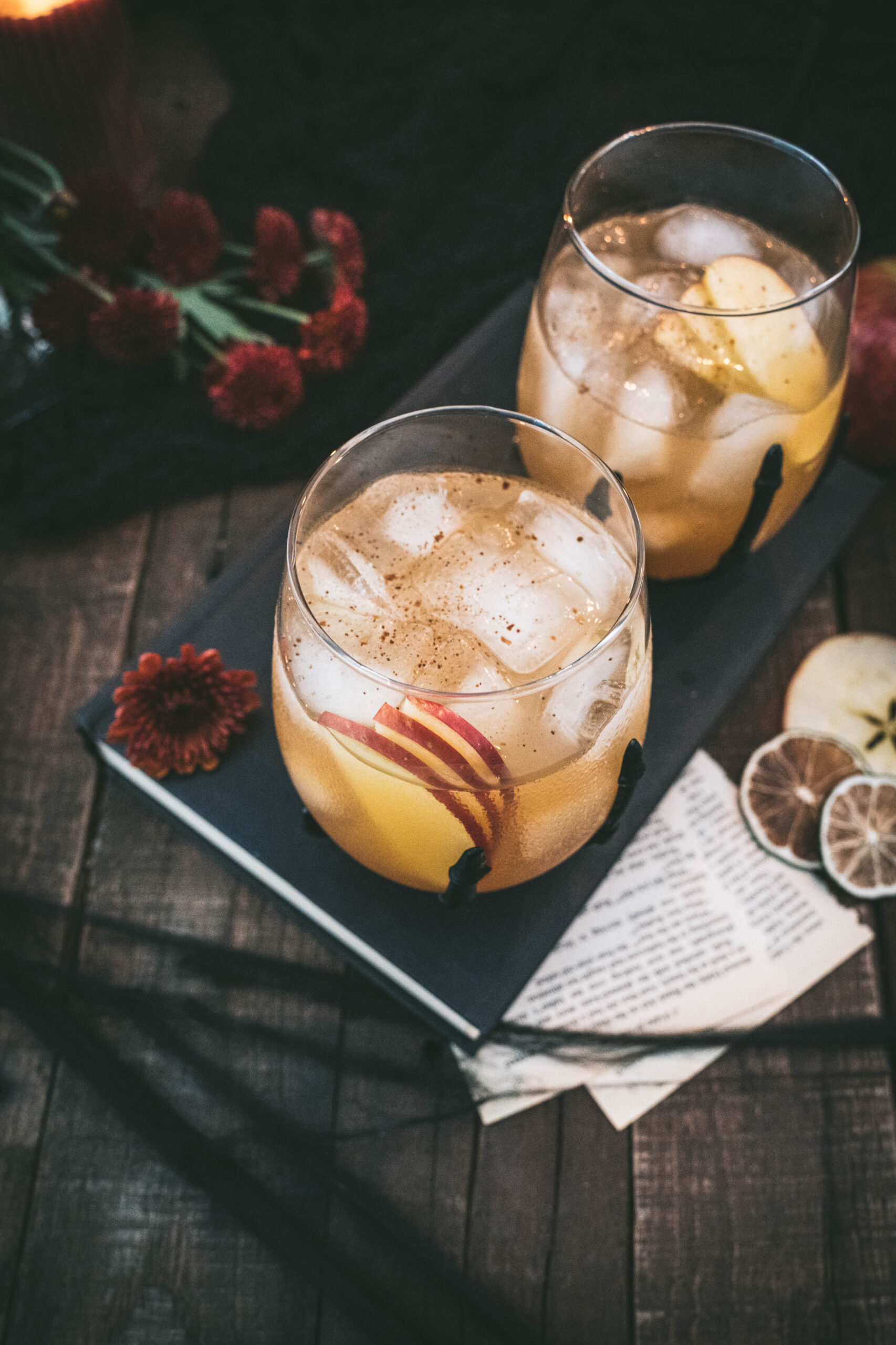 chai apple cider cocktail in skeleton cocktail glasses on wood surface, surrounded by red mum flowers, apple slice and dried limes