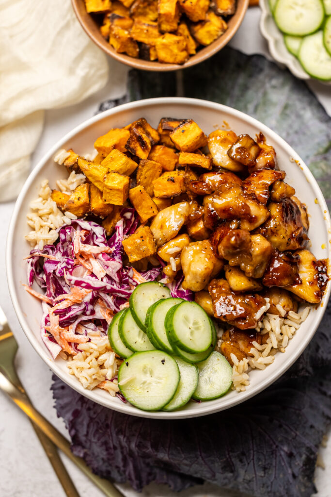 hot honey chicken bowl with cucumbers and sweet potatoes in a white bowl with a blue napkin underneath. Two gold forks are on the table on the left hand side.