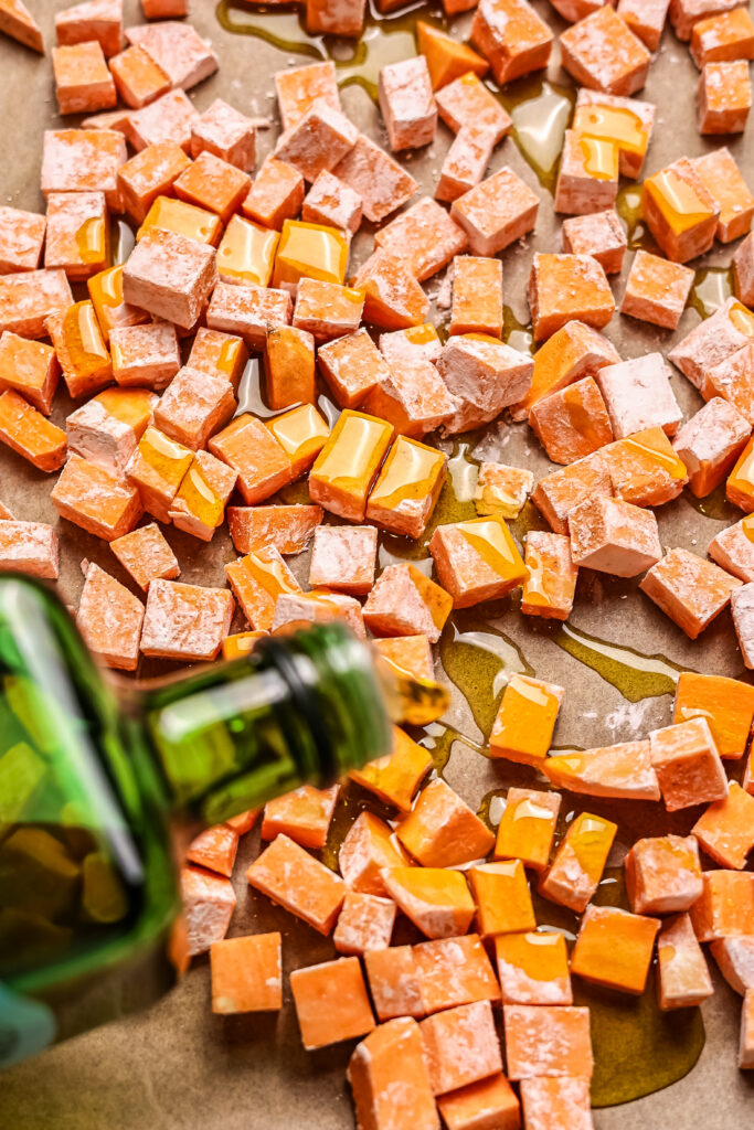 olive oil being poured over cubed swetet potatoes on baking sheet for hot honey chicken bowls