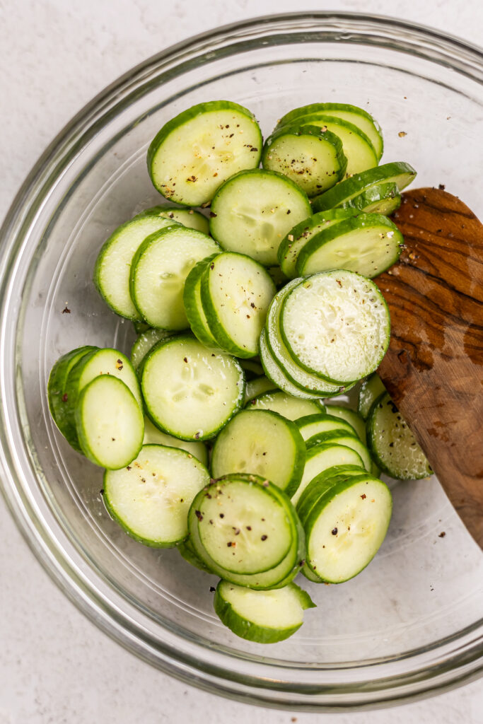 sliced cucumber seasoned with rice wine vinegar in bowl with wood spoon being prepped for hot honey chicken bowls