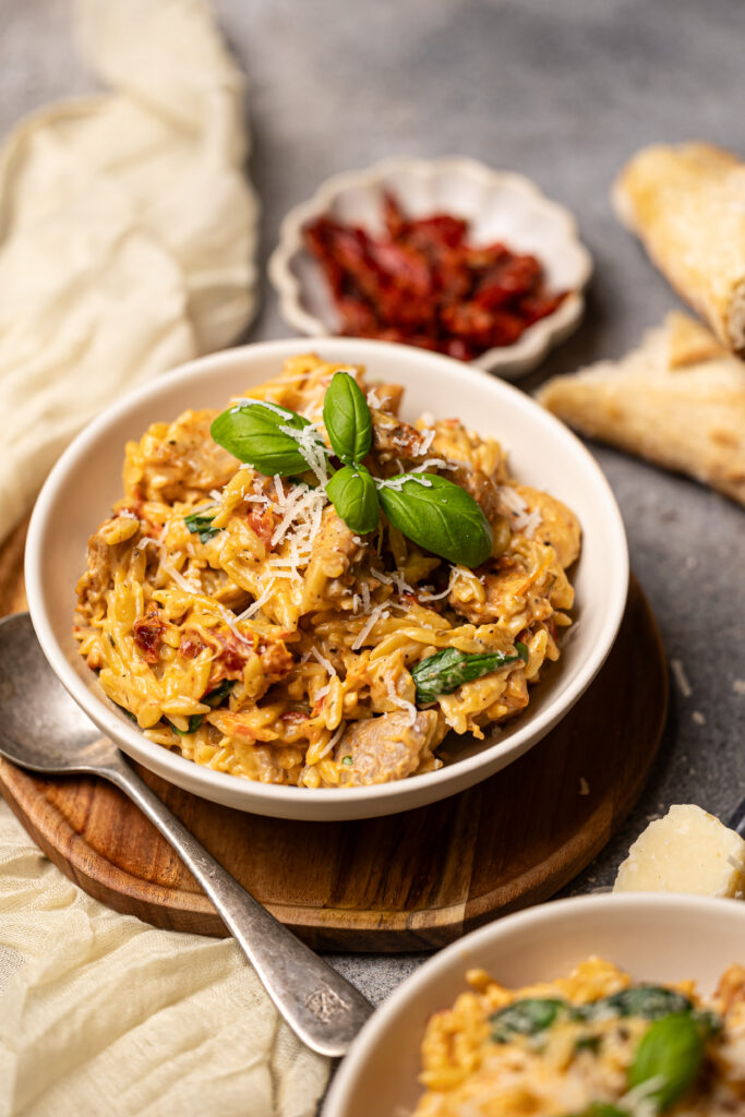 marry me chicken orzo in a bowl garnished with parmesan cheese and fresh basil. A cream napkin and bowl of sundried tomatoes is behind the main bowl.
