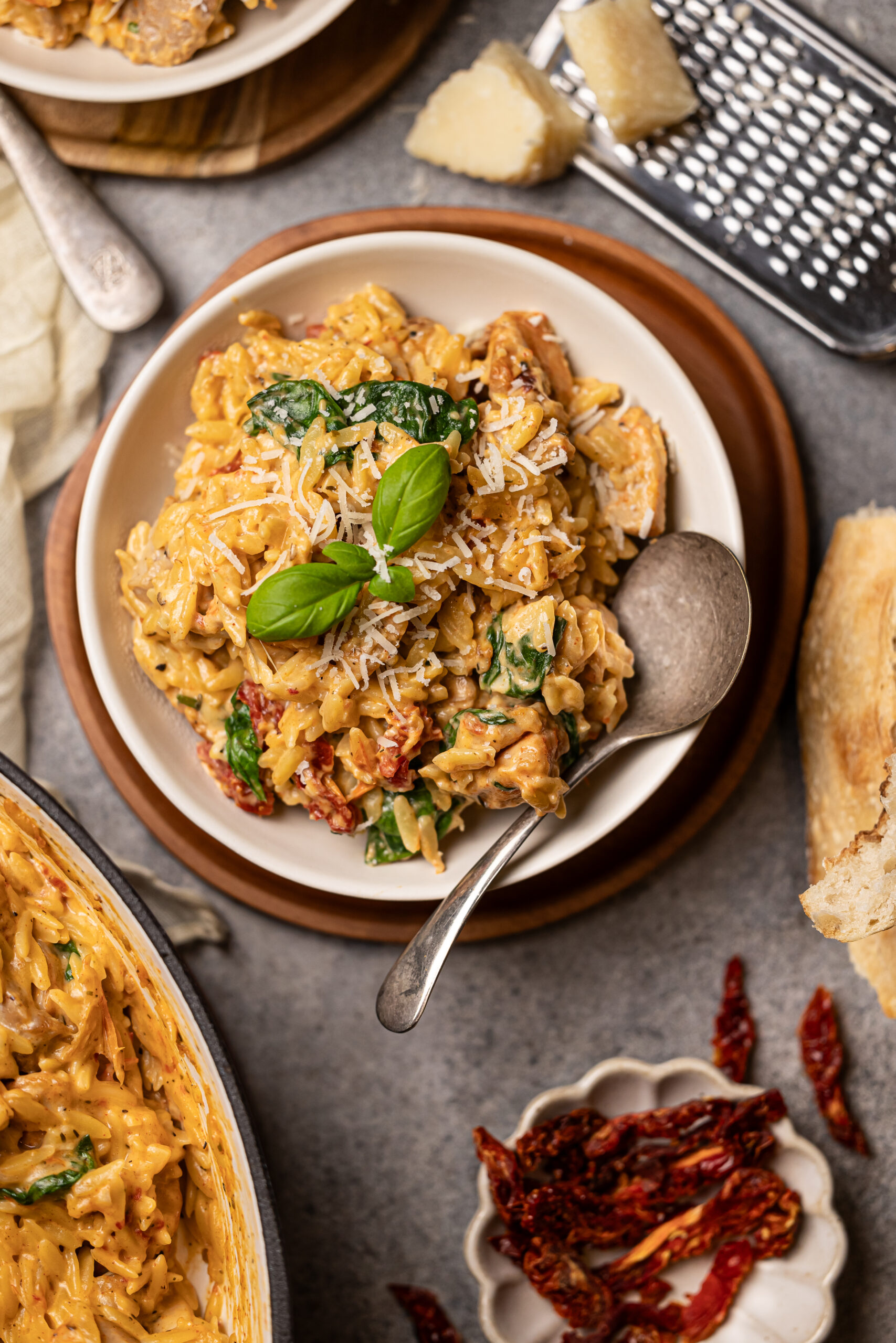 marry me chicken orzo in white bowl on top of wood charger with a silver spoon. Bread is to the right of the bowl and sundried tomatoes are in a small bowl below.