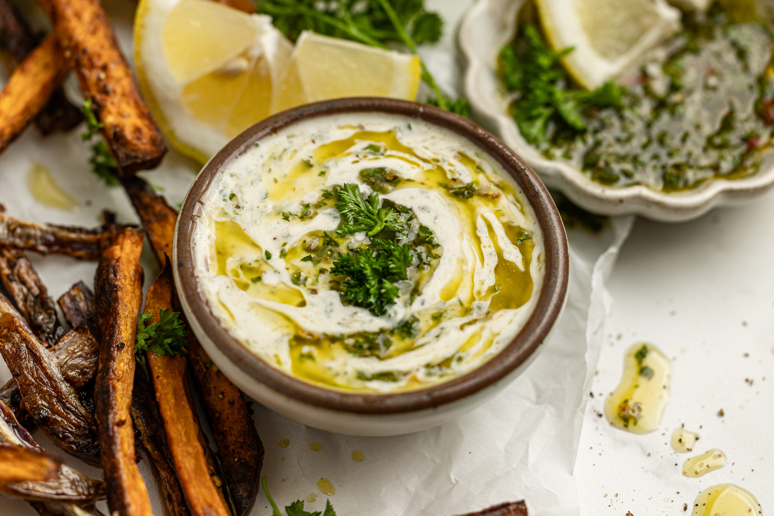 chimichurri aioli in a small bowl surrounded by crispy french fries, a lemon wedge and fresh parsley