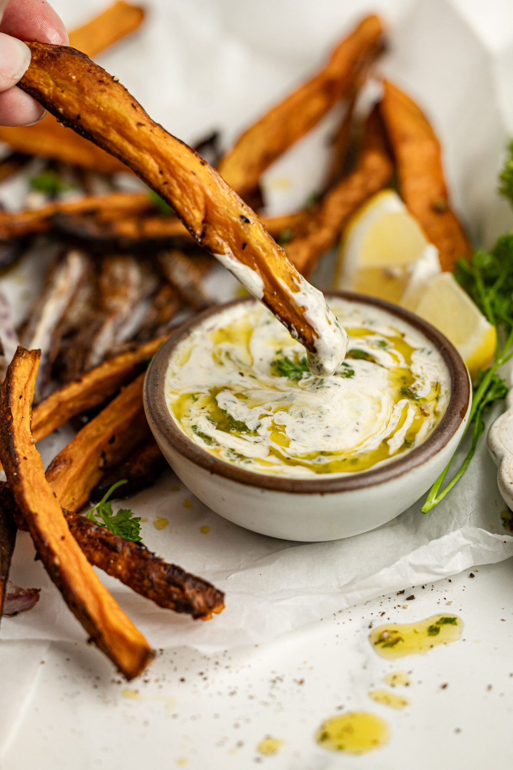 a side shot of a french fry being dipped in chimichurri aioli in a small bowl. The bowl is surrounded by crispy french fries, a lemon wedge and fresh parsley