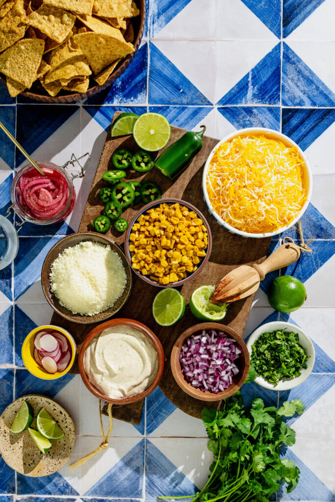 ingredient shot of elote nachos on a wood cutting board on a blue and white tile background
