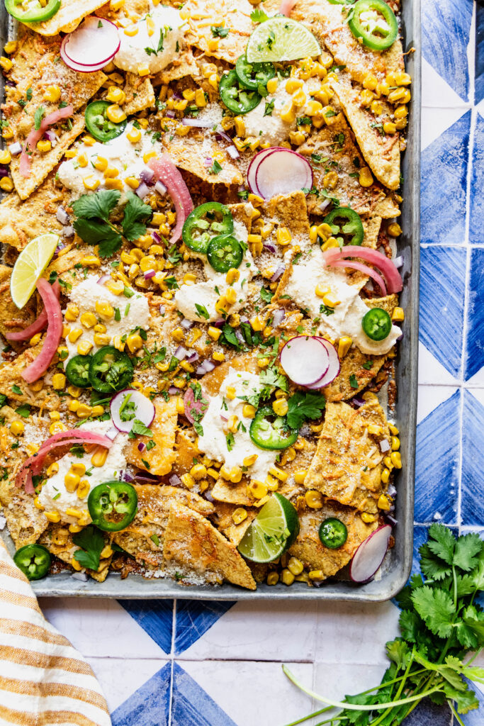 close up overhead shot of elote nachos garnished with jalapeno slices, cilantro, pickled onions and lime wedges. The nachos are on a sheet pan on a blue and white tile background