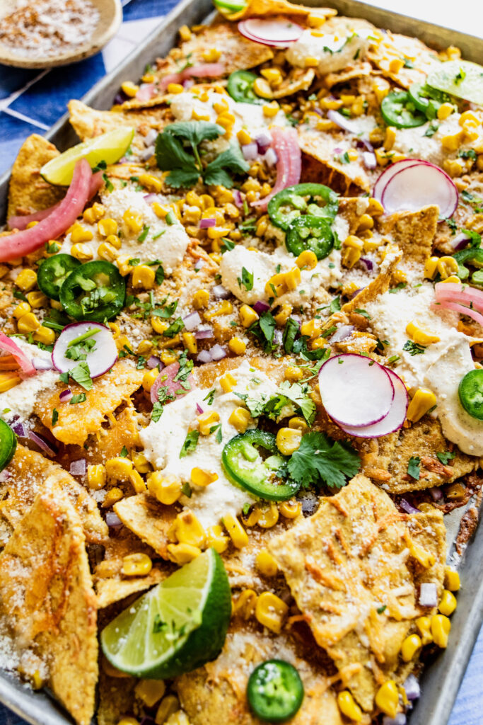 last two steps of elote nachos garnished with jalapeno slices, cilantro, pickled onions and lime wedges. The nachos are on a sheet pan on a blue and white tile background