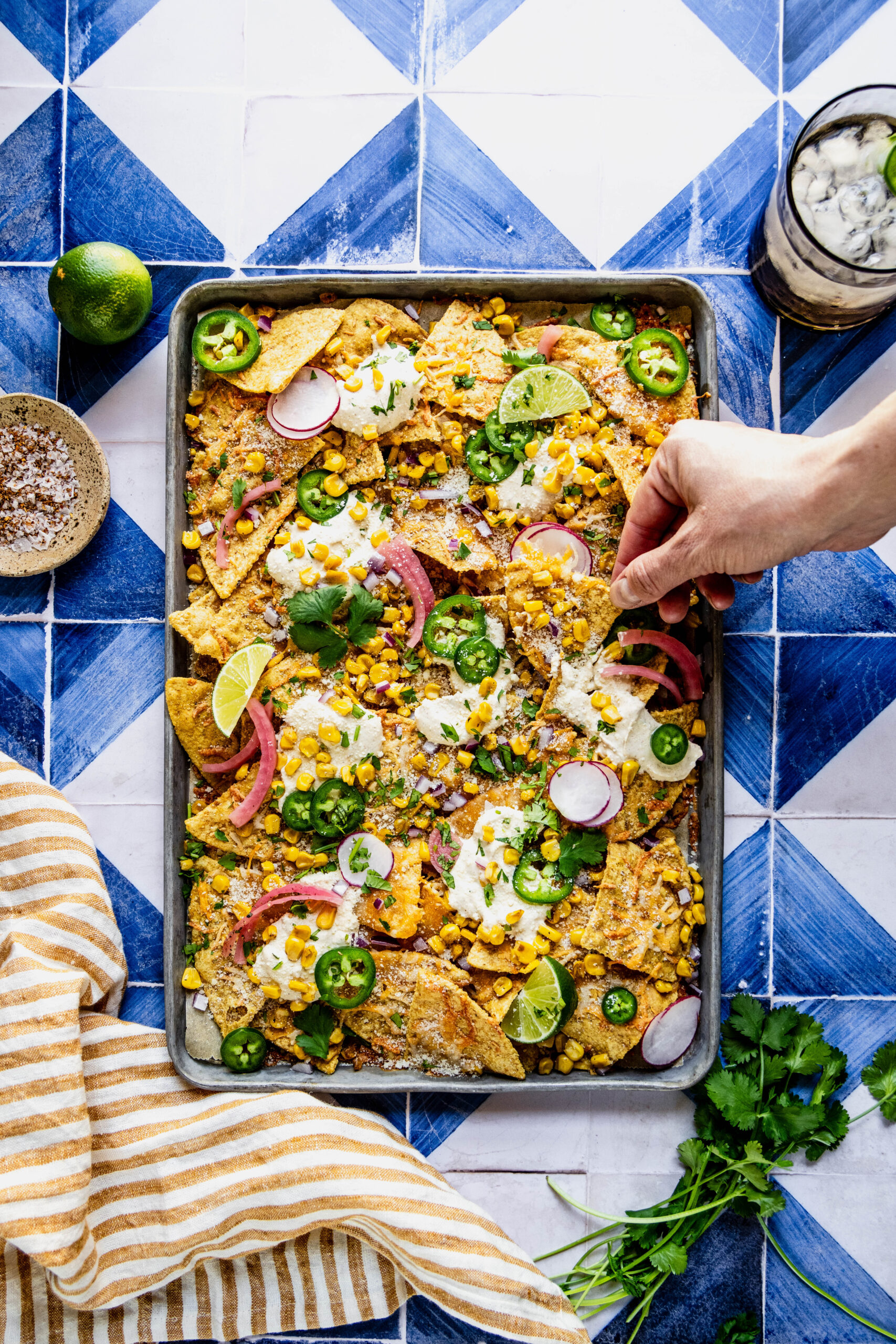 overhead shot of a hand reaching for a chip on the elote nachos garnished with jalapeno slices, cilantro, pickled onions and lime wedges. The nachos are on a sheet pan on a blue and white tile background