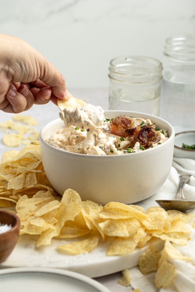 dipping potato chips into a bowl of homemade sour cream and onion dip on a white marble platter