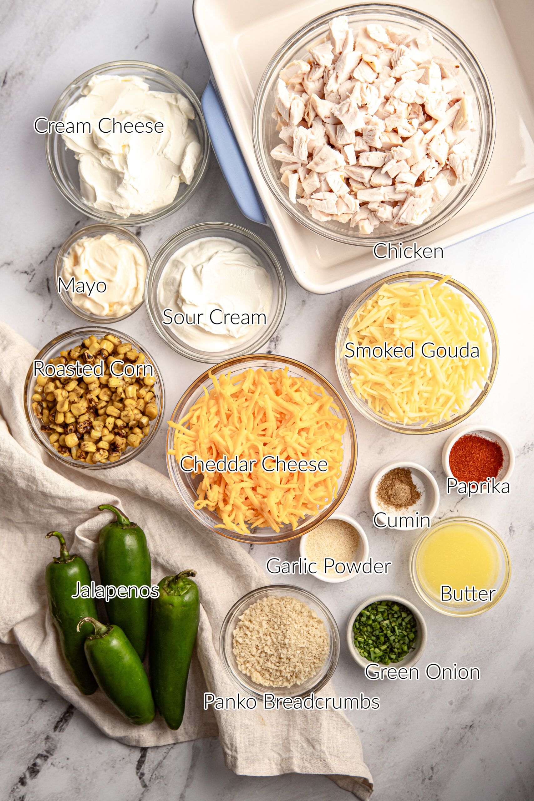 Overhead image of ingredients arranged on a marble surface for making jalapeño chicken popper dip, including fresh jalapeños, shredded chicken, cream cheese, sour cream, mayonnaise, roasted corn, shredded cheddar cheese, smoked gouda, garlic powder, smoked paprika, cumin, butter, green onion, and panko breadcrumbs.