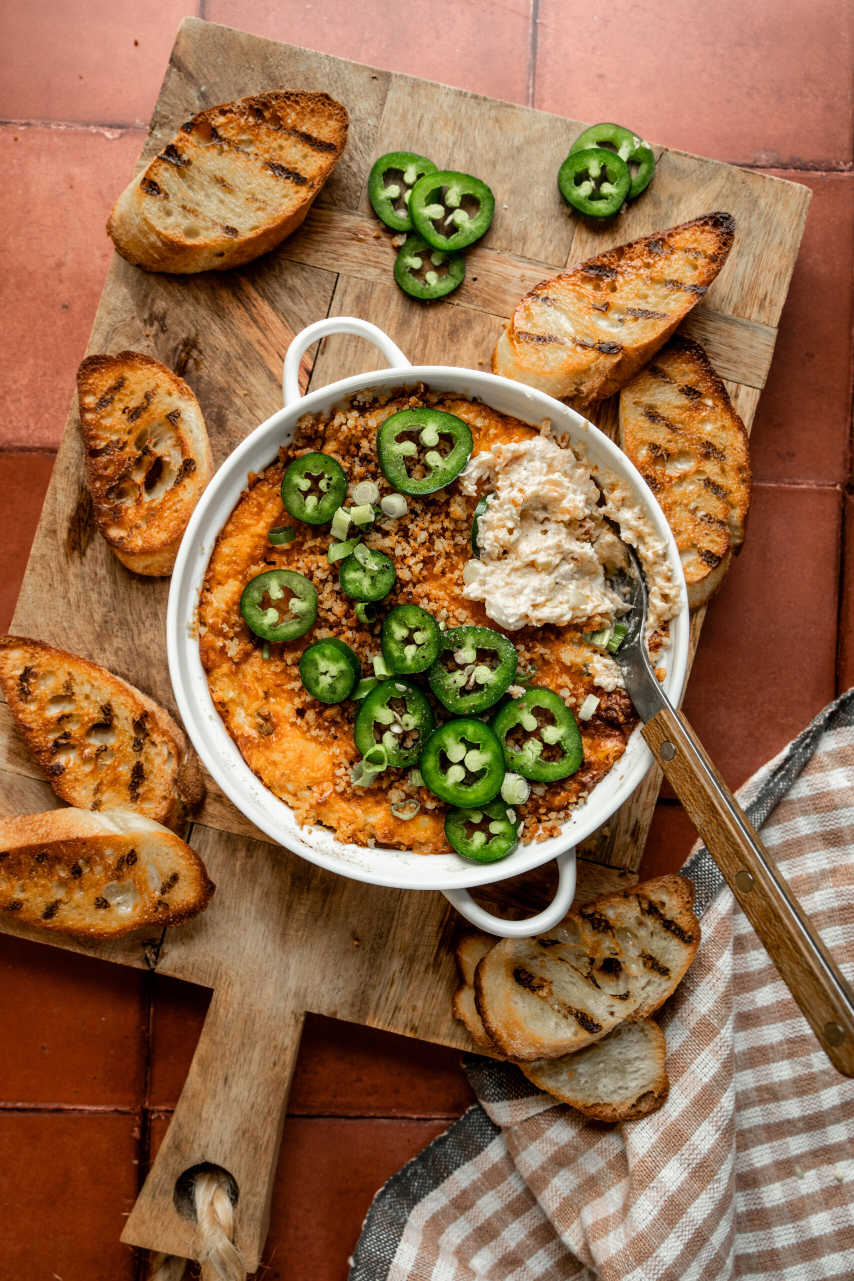 jalapenochxpopperdip 4Alt text: Overhead view of jalapeño chicken popper dip in a white baking dish, topped with sliced jalapeños, panko breadcrumbs, and green onions. A spoonful has been scooped out, showing the creamy texture. Surrounded by grilled crostini on a wooden serving board over a terracotta tile background.