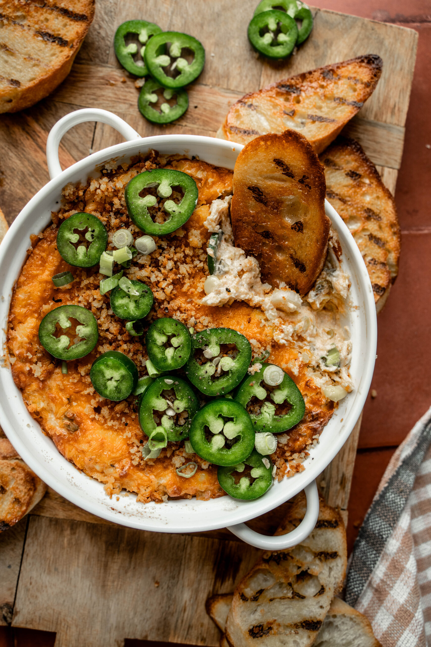 Close-up overhead image of jalapeño chicken popper dip in a white baking dish, topped with sliced fresh jalapeños, panko breadcrumbs, and green onions. A piece of grilled crostini is dipped into the creamy, cheesy dip, surrounded by more crostini on a wooden board.