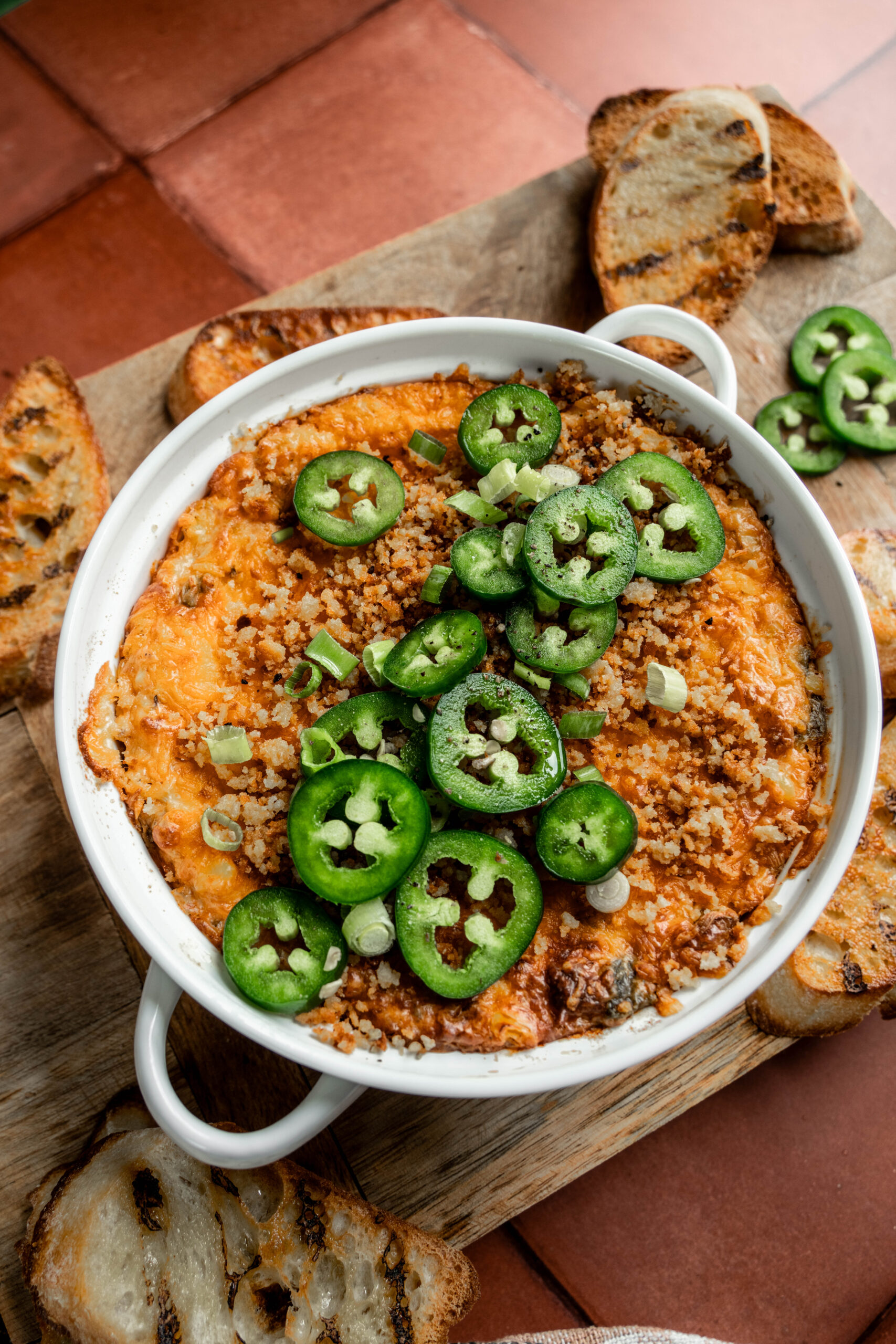 Alt text: Close-up of a bubbling hot jalapeño chicken popper dip in a white round baking dish, topped with sliced fresh jalapeños, golden panko breadcrumbs, and green onions, surrounded by grilled crostini on a wooden board.