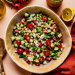 **Alt Text:** Overhead view of a fresh cucumber, tomato, and feta salad served in a large beige bowl on a peach-colored tiled surface. The salad is surrounded by ingredients including whole and sliced cucumbers, cherry tomatoes on the vine, a halved lemon, a bottle of olive oil, and a glass of water. A red cloth napkin and wooden serving utensils complete the scene, evoking a bright and summery Mediterranean vibe.
