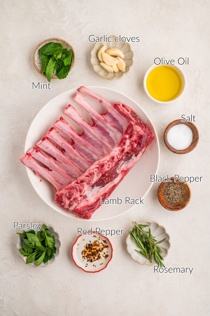 Overhead view of labeled ingredients for herb crusted rack of lamb recipe, including a raw lamb rack on a white plate, fresh mint, garlic cloves, olive oil, salt, black pepper, parsley, red pepper flakes, and rosemary arranged on a light surface.