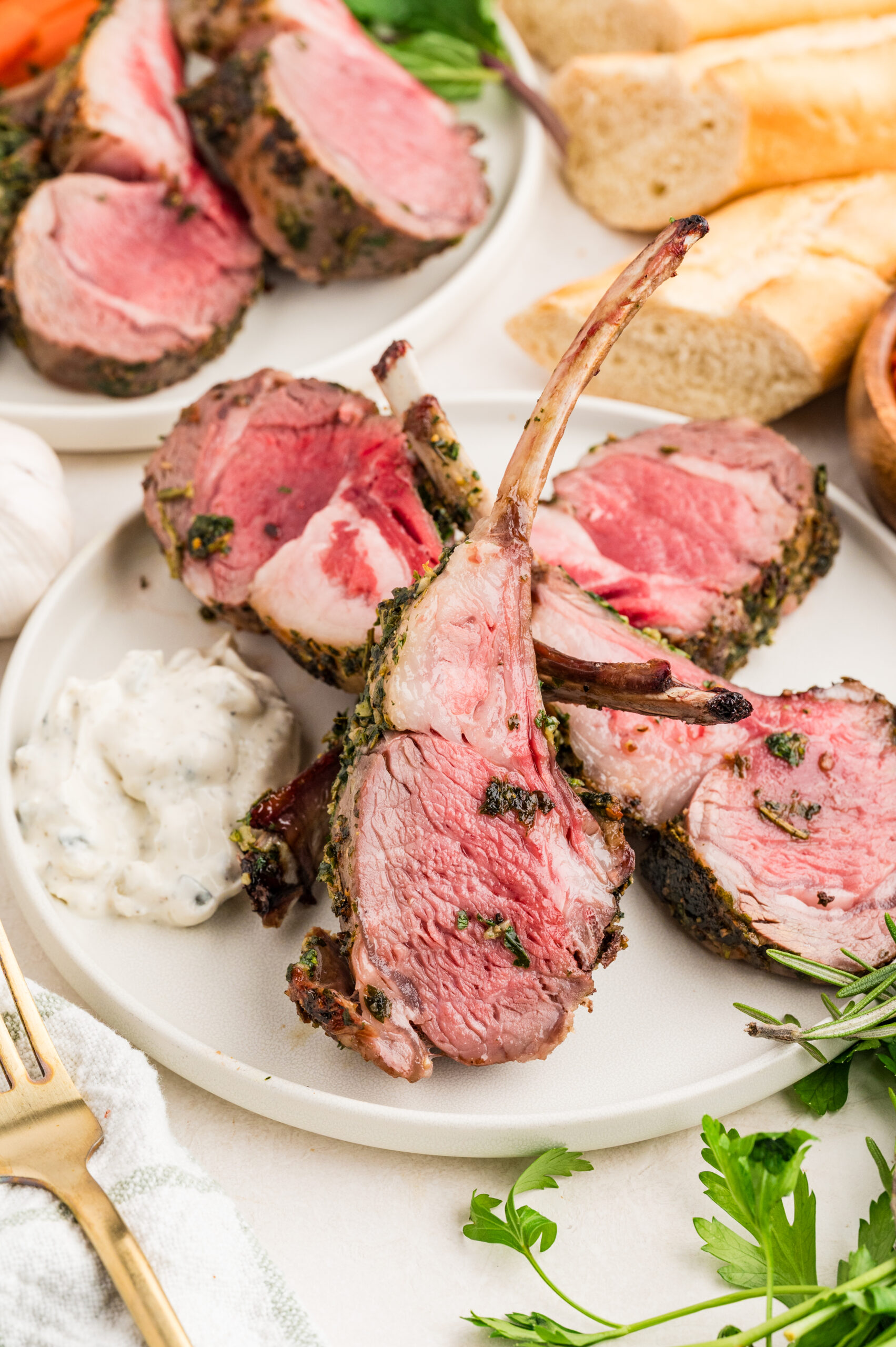 Sliced herb crusted rack of lamb recipe served on a white plate with a dollop of tzatziki sauce, garnished with fresh parsley and rosemary. Baguette slices and additional lamb pieces are visible in the background.