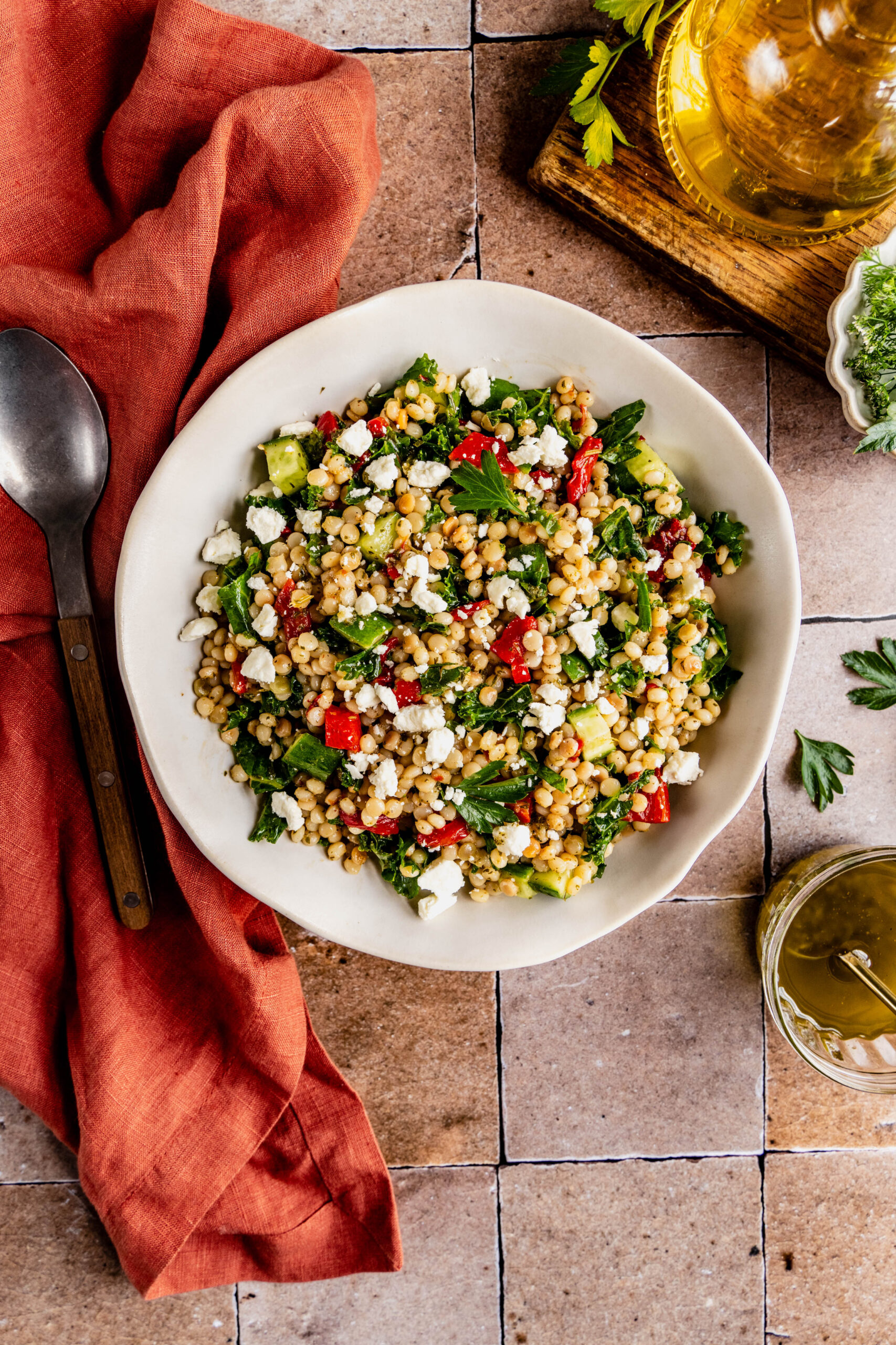overhead shot of a vibrant couscous kale salad served in a white bowl with a wooden-handled fork, featuring fluffy couscous, chopped kale, diced cucumbers, roasted red peppers, and crumbled feta cheese. The scene includes a small bowl of fresh herbs, a jar of olive oil, all styled on a rustic tiled surface with a burnt orange linen napkin.