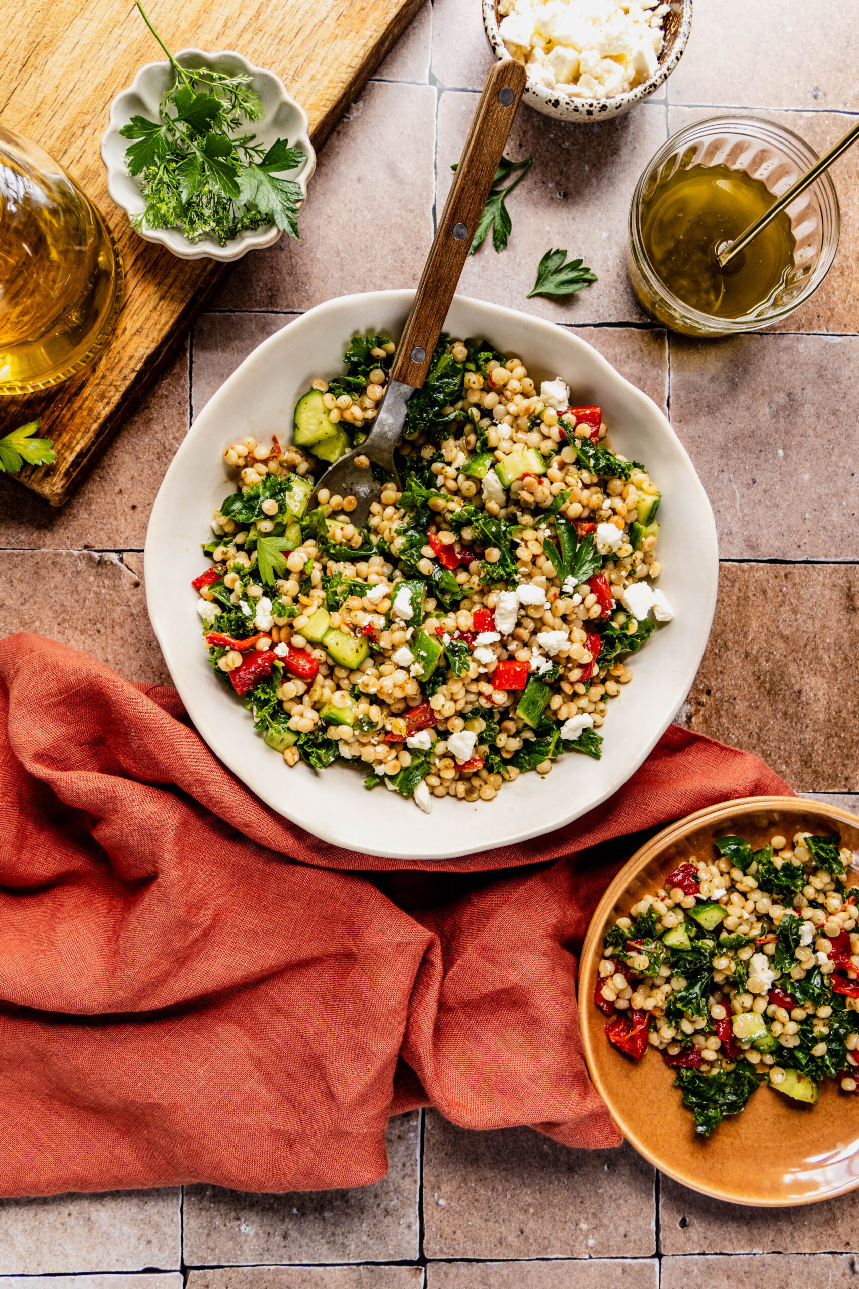 Overhead view of a vibrant couscous kale salad served in a white bowl with a wooden-handled fork, featuring fluffy couscous, chopped kale, diced cucumbers, roasted red peppers, and crumbled feta cheese. The scene includes a small bowl of fresh herbs, a jar of dressing, and a smaller serving of the salad in a brown bowl, all styled on a rustic tiled surface with a burnt orange linen napkin.