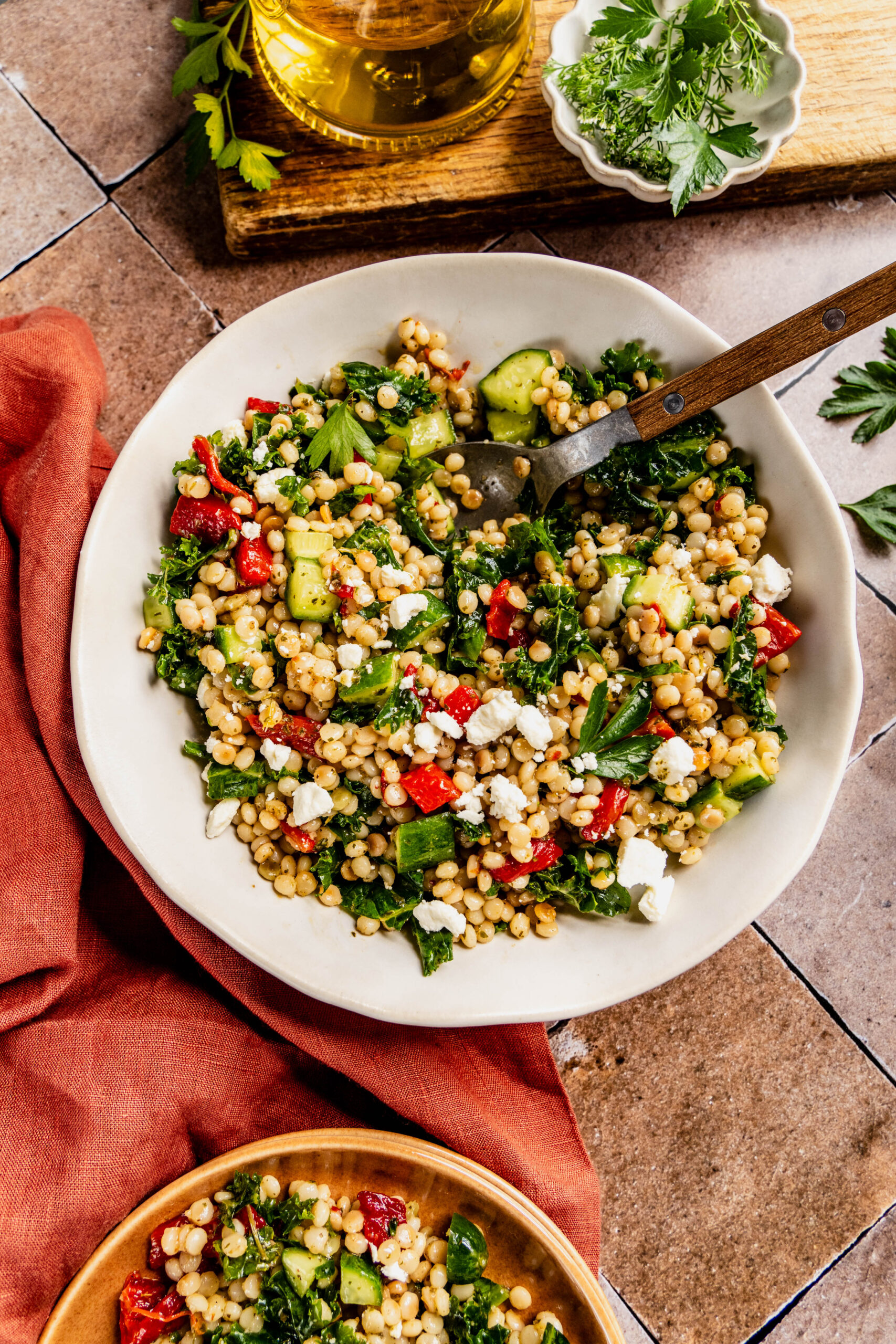close up view of a vibrant couscous kale salad served in a white bowl with a wooden-handled fork, featuring fluffy couscous, chopped kale, diced cucumbers, roasted red peppers, and crumbled feta cheese. The scene includes a small bowl of fresh herbs, a jar of olive oil, all styled on a rustic tiled surface with a burnt orange linen napkin.