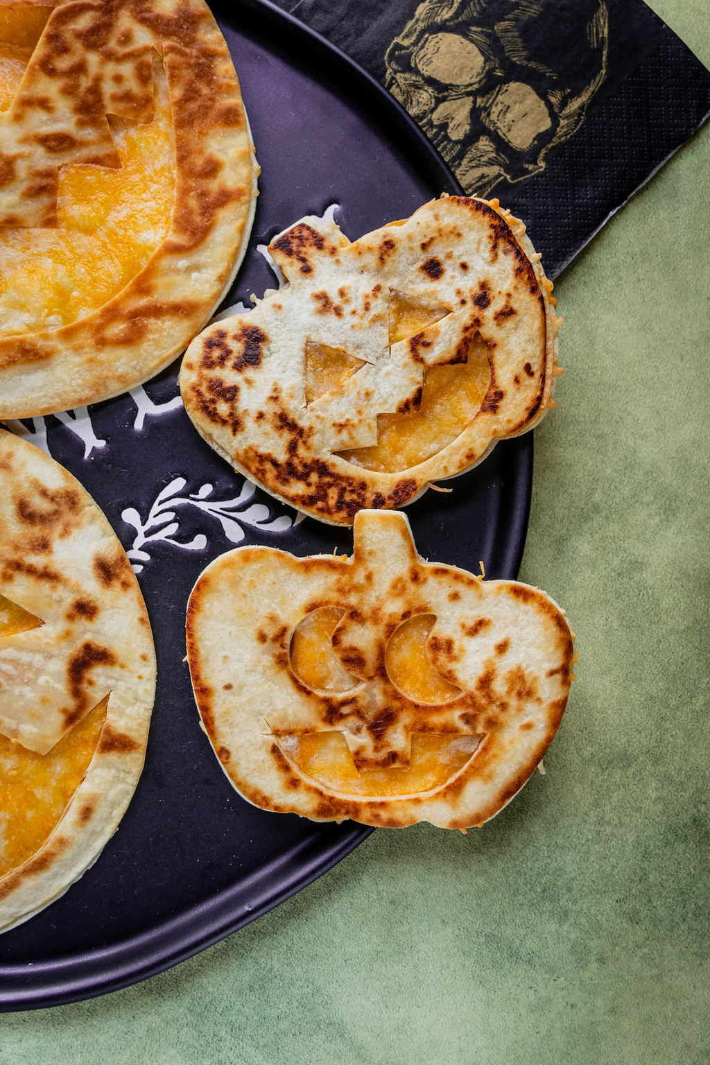Festive Halloween quesadillas shaped like jack-o’-lanterns, with melted cheese peeking through cut-out faces, served on a black plate with a side of salsa and decorative pumpkins in the background.