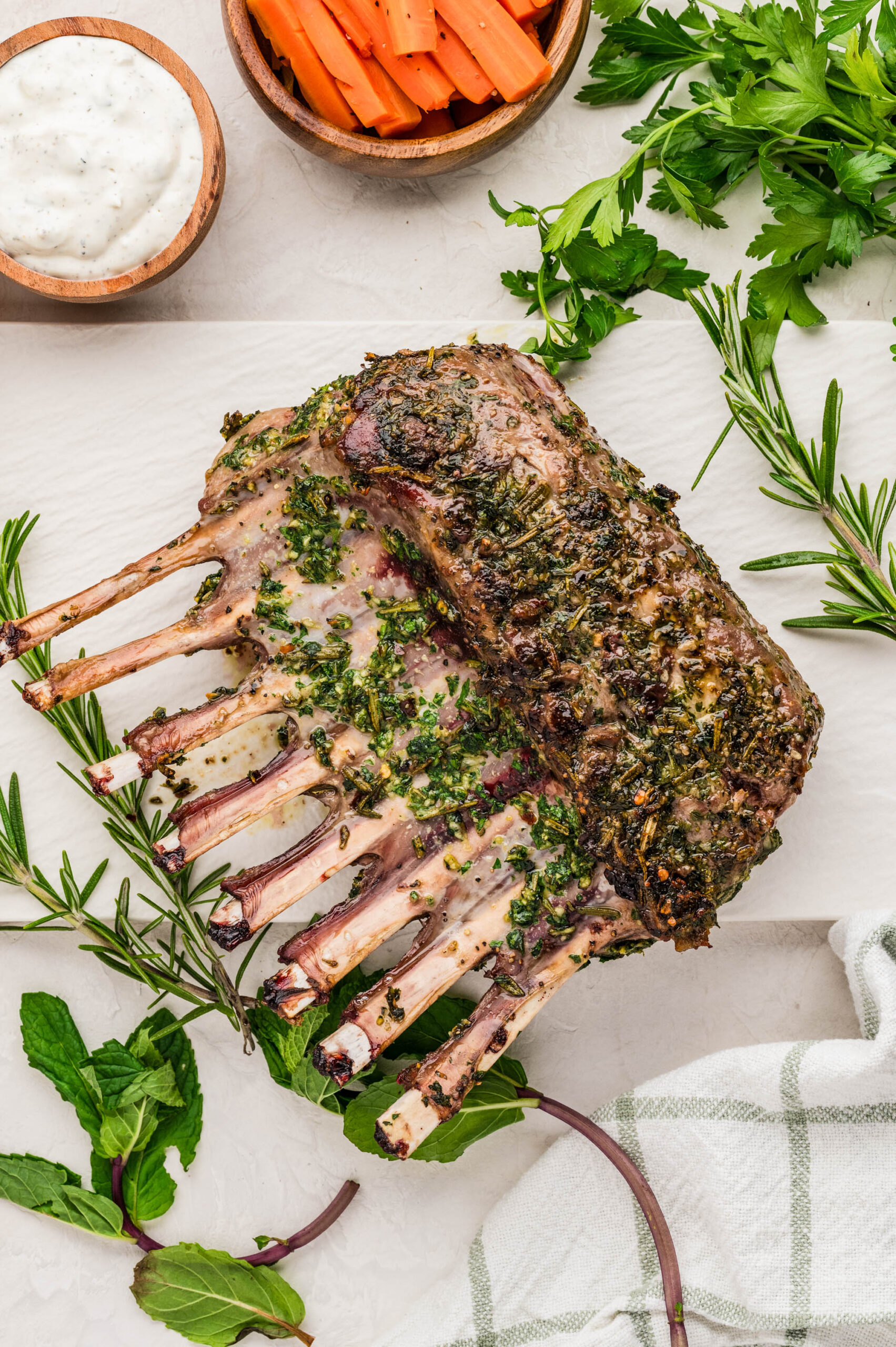 Alt text: Overhead view of herb crusted rack of lamb recipe on a white surface, garnished with fresh rosemary and mint. Served alongside carrot sticks in a wooden bowl, fresh parsley, and a small bowl of creamy tzatziki sauce.