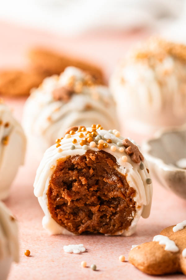 **Alt Text:**
Close-up of a **gingerbread truffle** coated in white chocolate and topped with festive gold and white sprinkles. The truffle has a bite taken out, revealing its rich, spiced gingerbread filling. More truffles and gingersnap cookies are softly blurred in the background on a pink surface, creating a warm holiday feel.