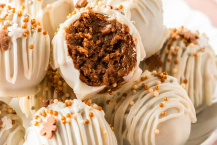 **Alt Text:** Close-up of homemade **gingerbread truffles** coated in smooth white chocolate, drizzled with extra white chocolate, and topped with gold and white festive sprinkles. One truffle is cut in half, revealing a rich, spiced gingerbread cookie filling inside. The truffles are arranged on a white plate with scattered sprinkles and small gingerbread cookie decorations, creating a cozy holiday dessert scene.