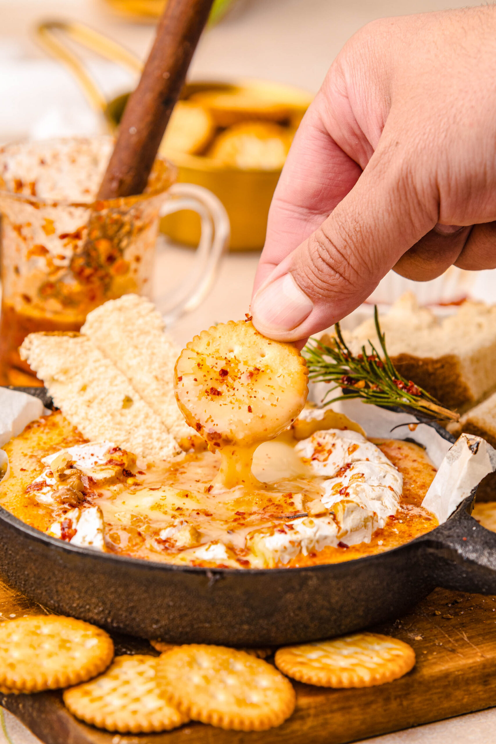 Close-up of a hand dipping a round cracker into warm, gooey baked Camembert topped with hot honey, red pepper flakes, and toasted walnuts. The melted cheese stretches beautifully from the skillet, surrounded by more crackers, bread slices, and a sprig of rosemary for garnish, illustrating the final serving of the baked Camembert recipe.