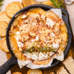 Overhead view of a baked Camembert recipe in a small cast-iron skillet, topped with bubbling hot honey, toasted walnuts, red pepper flakes, and a sprig of fresh rosemary. The melted cheese is surrounded by round buttery crackers and rustic bread slices for dipping, all arranged on a wooden serving board for a cozy, rustic appetizer presentation.