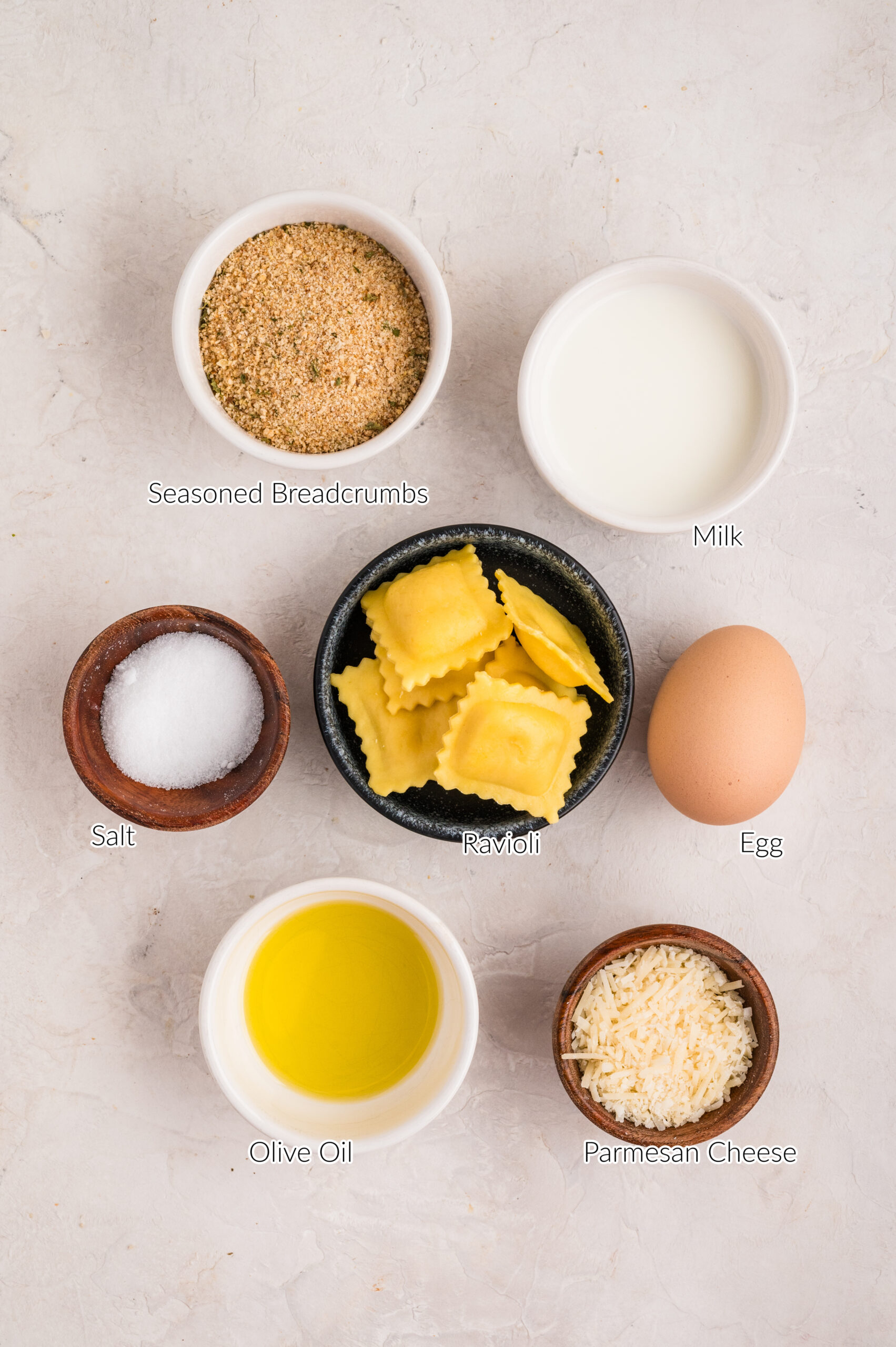Overhead view of ingredients for air fryer toasted ravioli, including a bowl of seasoned breadcrumbs, a small bowl of milk, a bowl of cheese ravioli, a whole egg, grated parmesan cheese, olive oil, and a small dish of salt arranged on a light textured surface.