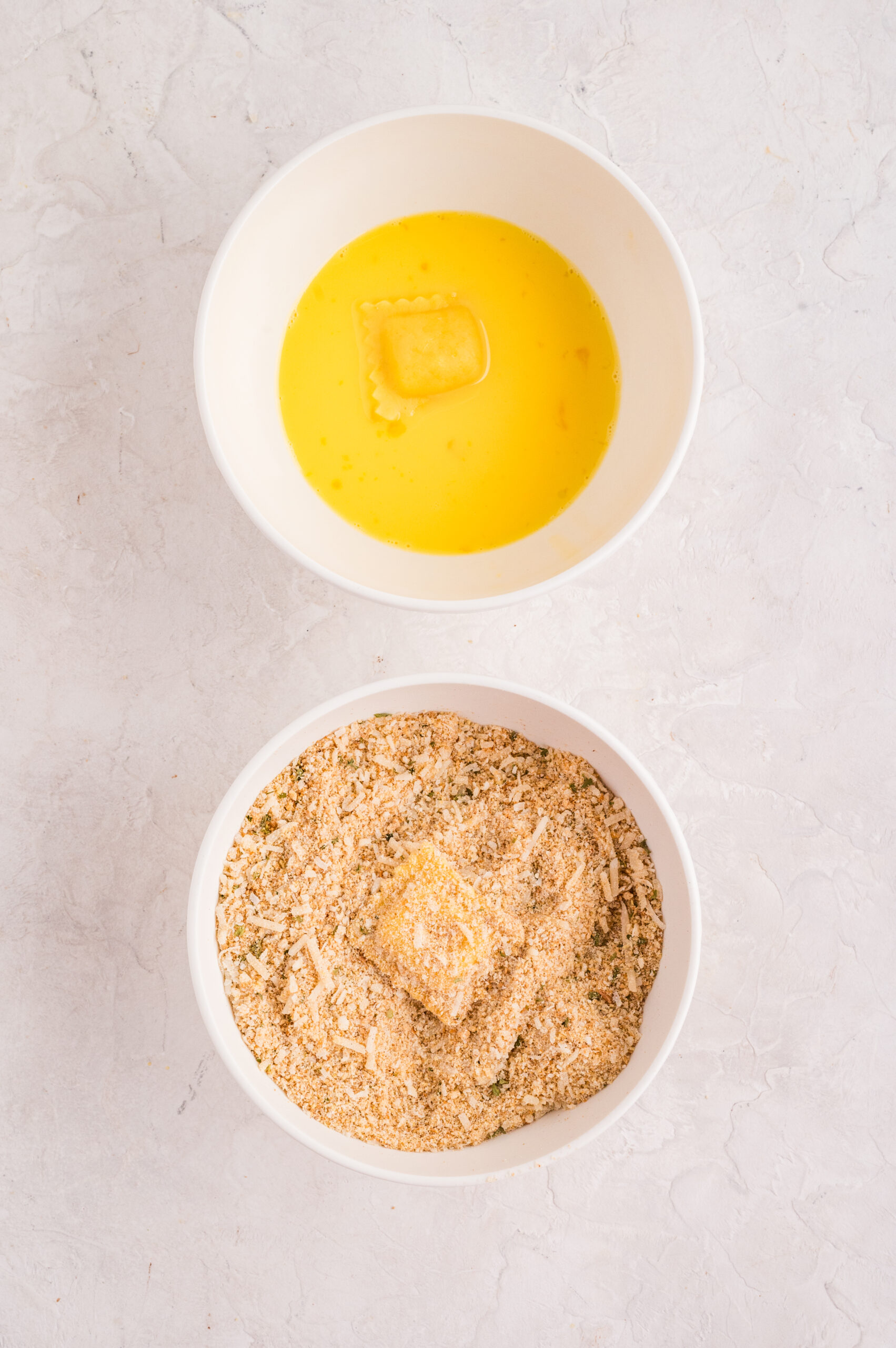 Overhead view of the breading process for air fryer toasted ravioli, showing one ravioli dipped in an egg mixture and another coated in seasoned breadcrumbs and parmesan cheese in a separate bowl.