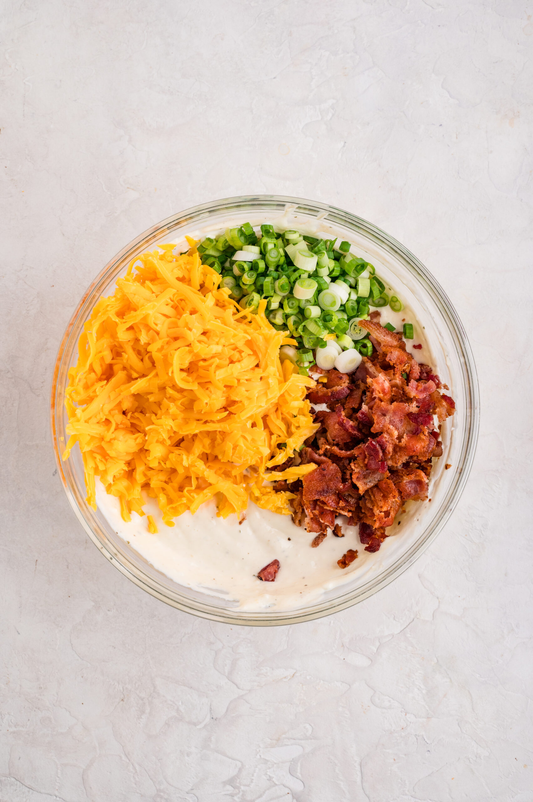 Glass mixing bowl filled with ingredients for bacon ranch cheddar dip, including shredded cheddar cheese, chopped green onions, and crispy bacon pieces on top of a creamy sour cream and mayo base, ready to be mixed together.
