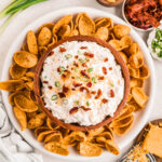 A wooden bowl filled with creamy bacon ranch cheddar dip topped with crispy bacon crumbles, shredded cheddar cheese, and chopped green onions, surrounded by scoops-style corn chips on a white platter. Small bowls of extra bacon, cheese, and green onions are arranged nearby for garnish.